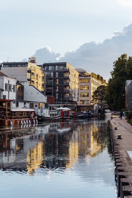 A serene riverside garden scene along the Grand Union Canal, featuring well-maintained grass lawns and garden beds bordered with trimmed hedges and small trees. The pathway runs parallel to the water,