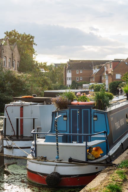 A serene riverside garden scene along the Grand Union Canal, featuring well-maintained grass lawns and garden beds bordered with trimmed hedges and small trees. The pathway runs parallel to the water,