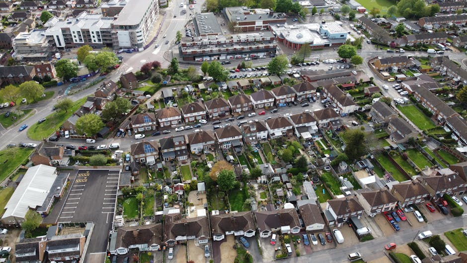 An aerial view of a suburban neighbourhood showing a mix of residential houses with small gardens, neatly maintained lawns, and flowerbeds. The gardens feature various plants, shrubs, and trees, with 